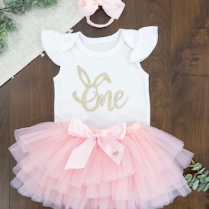 May include: A pink tulle tutu skirt with a pink satin bow and a white onesie with gold glitter lettering that says "one" and a bunny ear design. The onesie has short ruffle sleeves.
