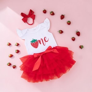 May include: A white baby bodysuit with a red strawberry graphic and the word "one" in red script. The outfit includes a red tulle tutu skirt, a red bow headband, and scattered strawberry decorations. The outfit is designed for a baby's first birthday.