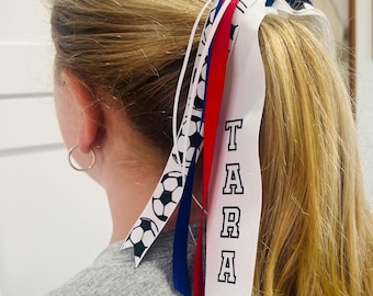 May include: A ponytail holder with red, white, and blue ribbons. One ribbon has the name "TARA" in black block letters. Another ribbon has a soccer ball pattern. The hair accessory is worn in a blonde ponytail.