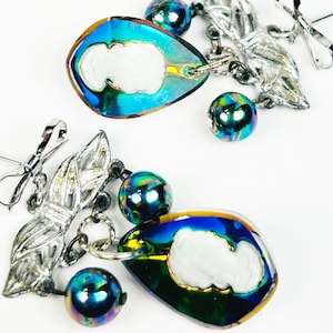 Peut inclure: Une paire de boucles d'oreilles pendantes bleu et vert irisé avec des accents argentés. Chaque boucle d'oreille présente un pendentif en forme de larme avec un motif de camée blanc, une feuille d'argent et trois perles bleu et vert irisé.