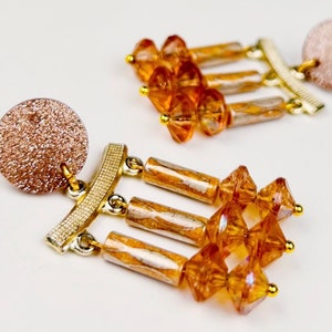 May include: A pair of gold and amber dangle earrings with a round, sparkly, pink base.
