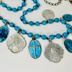Peut inclure: Un collier de perles bleu turquoise avec des médaillons religieux argentés et bleus. Le collier présente une variété de figures et de symboles religieux, notamment une croix, un cœur et une Vierge Marie.