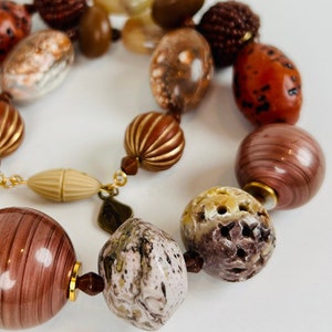 May include: A brown and white beaded necklace with a variety of textures and shapes. The necklace features a mix of round, oval, and irregular shaped beads. Some beads have a smooth finish, while others have a rough or textured surface. The necklace is made with a variety of materials, including wood, stone, and glass.