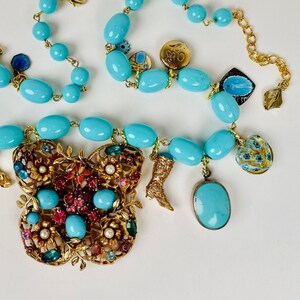 Op de afbeelding: Een turquoise en gouden ketting met een grote bloemvormige hanger. De ketting is gemaakt van turquoise kralen en heeft een verscheidenheid aan bedels, waaronder een laars, een bloem en een kleine vogel.