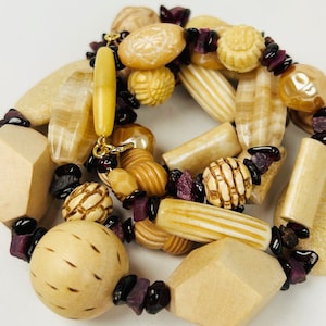 May include: A multi-strand necklace made of natural wood beads in various shapes and sizes, including round, oval, and rectangular. The beads are a light beige color and are interspersed with small, dark purple beads.