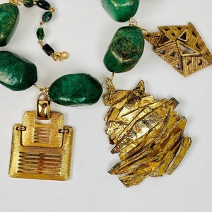 Puede incluir: Un collar de declaración dorado y verde con piedras verdes grandes, eslabones de cadena dorados y varios colgantes de amuleto dorados.