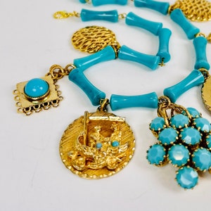 May include: A turquoise and gold necklace with multiple charms. The necklace is made of turquoise plastic beads and features charms of grapes, a chariot, and a figure of a woman.