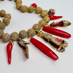 Può includere: Una collana d'oro con ciondoli rossi e dorati. La collana è realizzata con piccole perle dorate e ha una varietà di ciondoli, tra cui una corona, una scarpa e un peperoncino.
