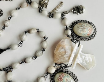 May include: A white beaded necklace with multiple charms, including a white shell, a small metal cage, a white flower, a white and black patterned charm, and a white and black patterned pendant.