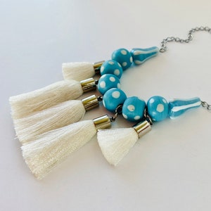 Puede incluir: Un collar de cuentas azules y blancas con borlas blancas. Las cuentas son azules con lunares blancos y las borlas están hechas de hilo blanco.