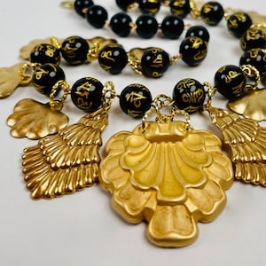 Puede incluir: Un collar de cuentas negras con dijes dorados en forma de conchas. Las cuentas están inscritas con letras negras.