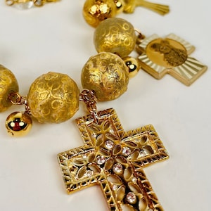 Peut inclure: Un collier en or avec un grand pendentif en forme de croix orné. La croix est en métal et présente un motif en filigrane. Le collier comporte également plusieurs autres breloques, notamment de petites perles, une cloche et une croix avec une image religieuse.