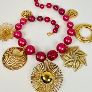 Peut inclure: Un collier de perles roses avec des breloques dorées, dont une fleur, un soleil, une coquille et un motif de feuille.
