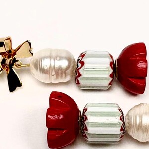 Puede incluir: Un par de pendientes de temática de caramelo rojo y blanco con acentos de lazo dorado. Cada pendiente presenta una cuenta roja en forma de caramelo, una cuenta rayada blanca y verde, una cuenta de perla blanca y un lazo dorado.