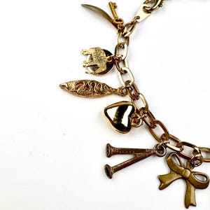 May include: A gold-toned charm bracelet with various charms, including a key, a sword, an elephant, a fish, a heart, a bow, and two trumpets. The charms are attached to a chain-link bracelet. The background is white.