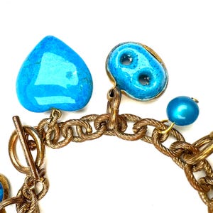 May include: Close-up of a gold-toned chain bracelet with turquoise heart and oval charms, and a blue bead. The bracelet has a toggle clasp. The charms are a bright turquoise color.