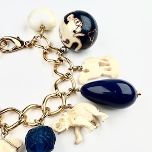 Peut inclure: Bracelet à breloques dorées avec divers charms. Les breloques comprennent un cœur blanc, un globe bleu foncé avec des accents blancs, des breloques éléphants ivoire et un charm en forme de goutte d'eau bleu foncé. Le bracelet a un fermoir mousqueton.