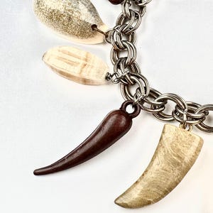 May include: A silver chain bracelet with multiple decorative charms. The charms include a dark brown horn-shaped pendant, a light beige curved pendant, and two other pendants with a beige and brown color scheme.