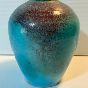 May include: A ceramic vase with a rounded body and a narrow neck. The vase features a gradient of colors, transitioning from a deep reddish-brown at the top to a vibrant turquoise blue at the base. The surface has a speckled texture.