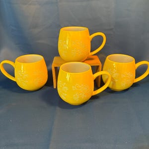 May include: Four yellow ceramic mugs with a floral design. The mugs have a rounded shape with a handle and a white interior. One mug is placed on a small wooden stand. The mugs are arranged on a blue surface.