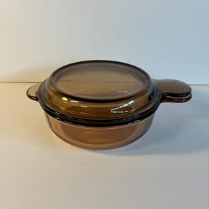 May include: A brown glass casserole dish with a matching lid. The dish has two small handles on either side and is designed for oven use. The glass is a warm, amber color, and the lid fits snugly on top.