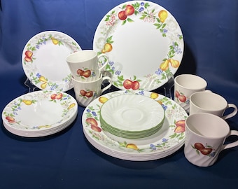 May include: A white ceramic dinnerware set featuring a colorful fruit pattern of red apples, yellow pears, and blue berries. The collection includes large dinner plates, smaller salad plates, saucers, and mugs, all decorated with green leaves and floral accents.