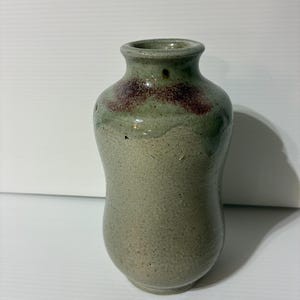May include: A small, handcrafted ceramic vase with a speckled, textured surface. The vase has a rounded body and a narrow neck. The top of the vase features a band of dark red and green glaze. The vase is approximately 5 inches tall.
