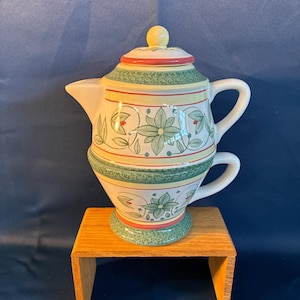 May include: A decorative ceramic teapot and teacup set. The teapot has a lid with a yellow knob and is decorated with green floral patterns, red accents, and yellow stripes. The teacup is stacked beneath the teapot. The set is displayed on a small wooden stand.