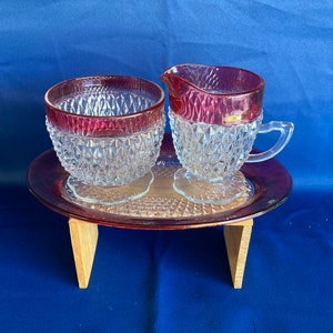 May include: A vintage glass sugar bowl and creamer set with a clear base and a pink rim. The set is on a matching pink tray with a diamond pattern.
