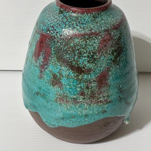 May include: A handcrafted ceramic vase with a unique, textured surface. The vase features a turquoise glaze over a brown base, with maroon accents. The opening is rimmed with maroon. The vase has a rounded shape and is suitable for home decor.