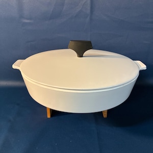 May include: A light gray, oval-shaped casserole dish with a matching lid and a black handle. The dish sits on a wooden stand. The overall design is simple and functional, suitable for cooking and serving.
