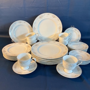 May include: A complete white porcelain dinnerware set with a floral design in shades of tan and light blue. The set includes plates, saucers, and teacups. The plates are stacked in neat piles, and the cups and saucers are arranged in groups.