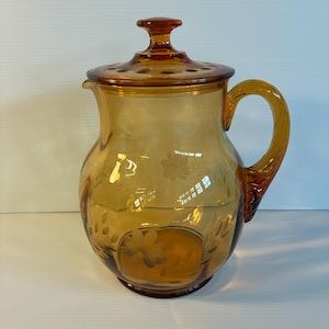 May include: Amber-colored glass pitcher with a lid and handle. The pitcher has a round body with etched floral designs. The lid has a small knob and a series of holes. The handle is curved and attached to the side of the pitcher.