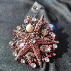 May include: A decorative brooch featuring a starfish design with pink and clear crystals and a large pearl. The brooch has a gold-tone metal base and a clear plastic clip.