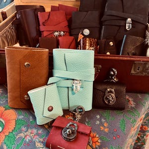 May include: A collection of handmade leather goods in various colors, including brown, red, black, and teal. The items include journals, wallets, and small pouches, some with decorative clasps and closures. The products are displayed on a patterned fabric.