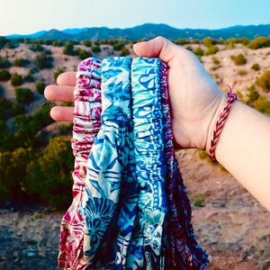 May include: Three patterned headbands in shades of blue and pink are held up against a backdrop of a mountain range. The headbands have a floral and geometric design. The background features a clear sky and a landscape of green bushes.