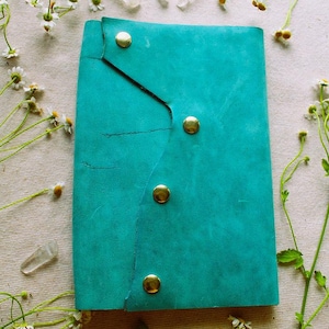 Puede incluir: Un diario de cuero turquesa con tres cierres de botón de latón. El diario está rodeado de flores blancas y cristales de cuarzo transparentes. La cubierta tiene una apariencia texturizada y una línea diagonal negra.