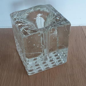 Pode incluir: Um castiçal quadrado de vidro transparente com uma superfície texturizada. O castiçal está sobre uma superfície de madeira. O castiçal foi projetado para conter uma única vela.