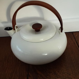Puede incluir: Hervidor de esmalte blanco con asa y pomo de tapa de madera marrón. El hervidor tiene un cuerpo redondeado y una boquilla con interior negro. El asa se arquea sobre la parte superior. El hervidor está sobre una superficie de madera oscura.