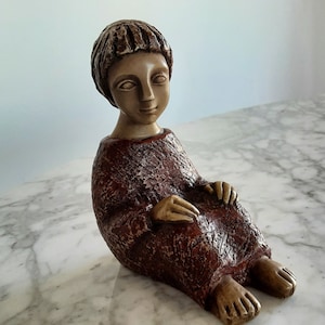 May include: A ceramic figurine of a person sitting with their legs crossed. The figurine is painted in shades of brown and red, with a textured surface.