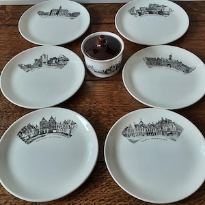May include: Set of six white ceramic plates with black and white illustrations of buildings. The plates are arranged in a circle around a brown ceramic sugar bowl.