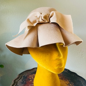 May include: A beige felt hat with a large bow on top. The hat is sitting on a yellow mannequin head.