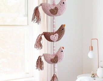 May include: A pink and brown crocheted bird mobile with three birds hanging from a string of wooden beads. The birds have pink and brown crocheted bodies with a pink heart on each bird's chest.