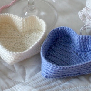 May include: Two crocheted heart-shaped baskets, one white and one light blue, are displayed on a white surface.