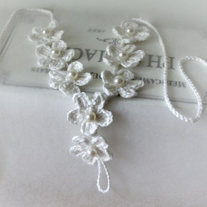 May include: White crocheted flower necklace with pearl accents. The necklace is made of delicate white yarn and features a series of crocheted flowers with a pearl bead in the center of each flower. The necklace is tied with a long white cord.