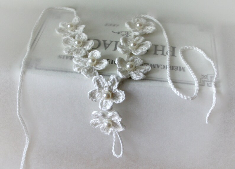 May include: White crocheted flower necklace with pearl accents. The necklace is made of delicate white yarn and features a series of crocheted flowers with a pearl bead in the center of each flower. The necklace is tied with a long white cord.