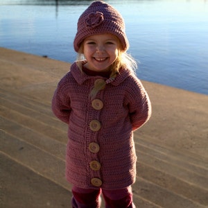 May include: A young person wearing a pink crocheted cardigan with brown buttons and a pink crocheted hat with a flower detail. The person is smiling and standing on a sandy beach.