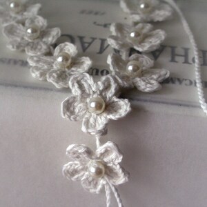 May include: White crocheted flower necklace with pearl accents. The necklace is made of delicate crocheted flowers with a pearl bead in the center of each flower. The necklace is strung on a white cord.