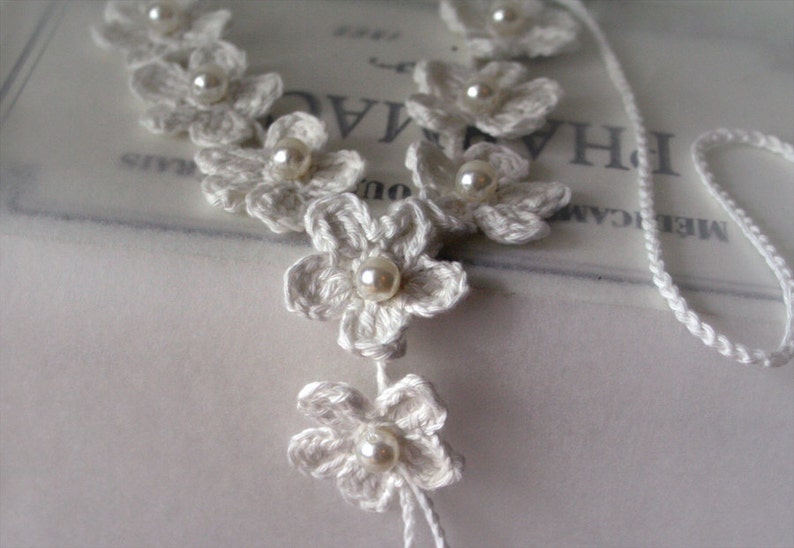 May include: White crocheted flower necklace with pearl accents. The necklace is made of delicate crocheted flowers with a pearl bead in the center of each flower. The necklace is strung on a white cord.