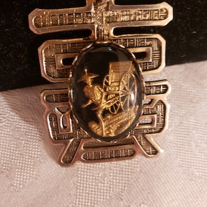 Peut inclure: Pendentif en or avec un camée ovale noir représentant une personne en or sur un chariot. Le pendentif est en forme de caractère chinois avec des détails complexes.